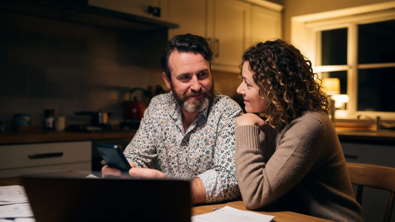 A couple at a kitchen table in the evening, looking at a phone together with a laptop open beside them.