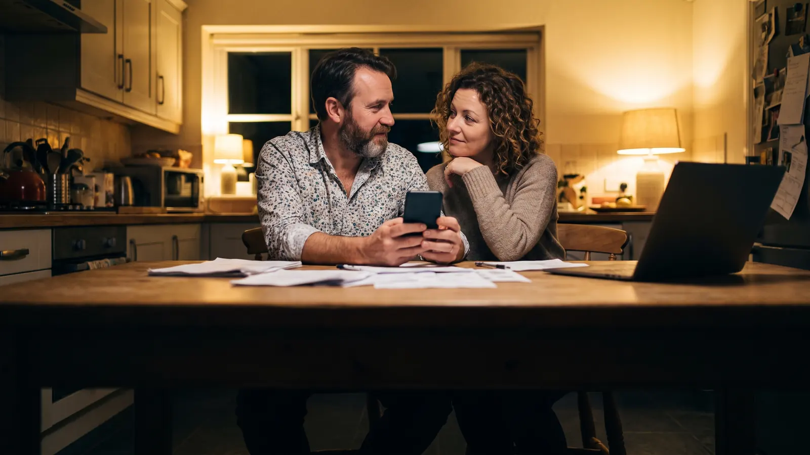 A couple at a kitchen table in the evening comparing something on a phone, papers and a laptop around them.