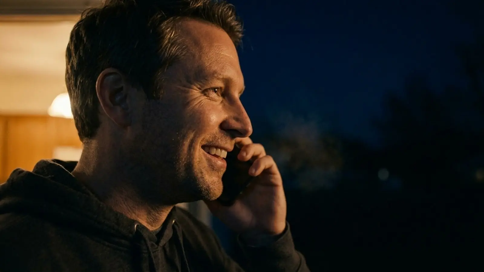 A woman sitting outdoors at dusk, holding a phone, seen from behind.